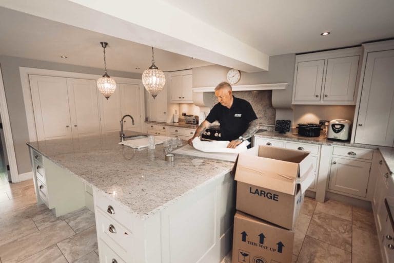 Man in a navy polo shirt wrapping glassware with packing paper on a large kitchen counter, with open cardboard boxes labeled 'LARGE' beside him.