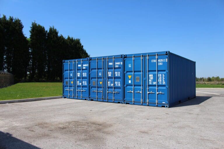 Row of large blue shipping containers placed on a paved outdoor area with grass and trees in the background under a clear blue sky.