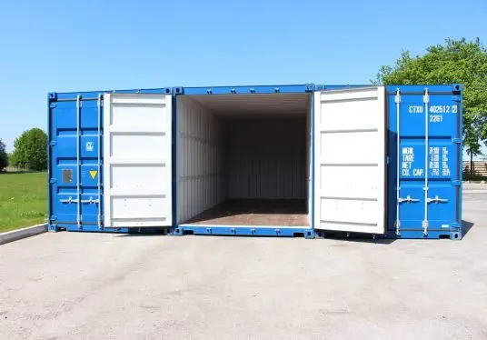 An empty blue shipping container with both front doors wide open, placed on a paved surface outdoors under a clear blue sky.