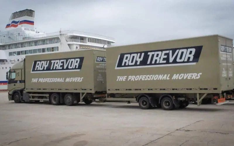 Two large beige moving trucks labeled 'Roy Trevor – The Professional Movers' are parked side by side near a dock, with a white passenger ferry in the background.