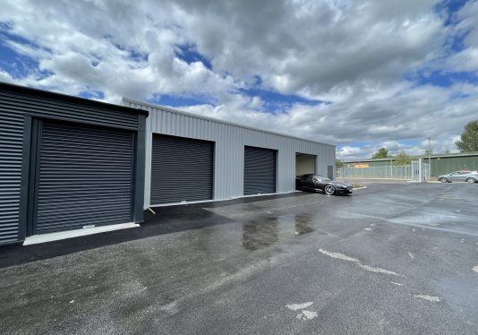 Exterior of Winsford storage facility with multiple grey roller door units, one open with a black car parked inside, under a cloudy sky.