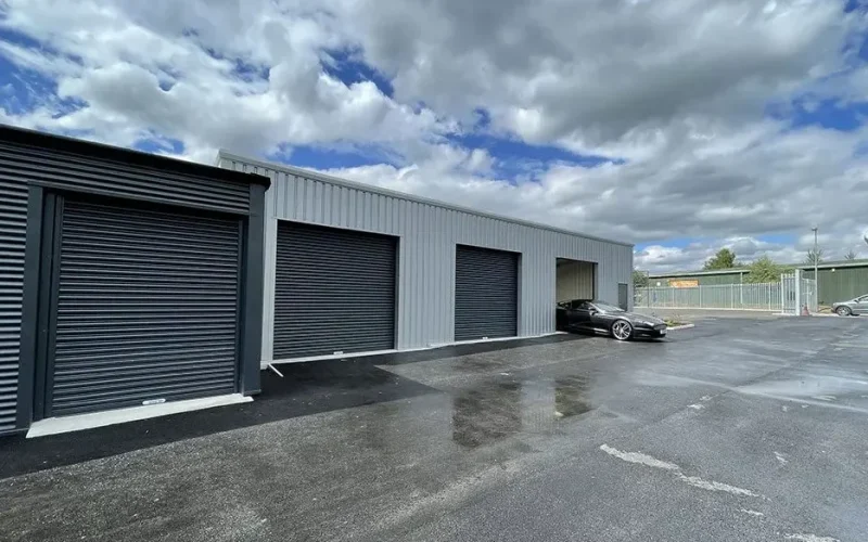 Outdoor self-storage units in Winsford with black roller doors, one unit open with a black car parked inside, under a cloudy sky.