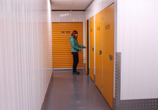 Woman in a green jacket unlocking a yellow self-storage unit door inside a brightly lit storage facility corridor.