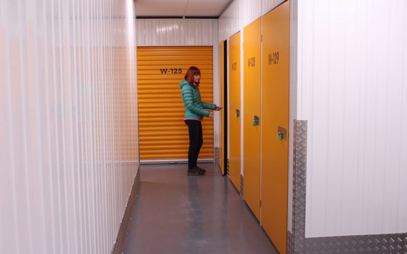 Woman in a green jacket unlocking a yellow self-storage unit door inside a brightly lit storage facility corridor.